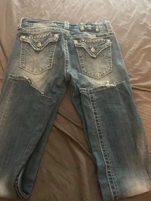 Blå miss me jeans med slitningar och nitar - Säljer ett par blå miss me jeans  med slitna detaljer och dekorativa nitar på fickorna. Jeansen har patchade partier och tydliga kontrastsömmar. Bakfickorna har lock och knappar, vilket ger en unik look. Perfekta för dig som gillar en edgy och trendig stil. Dem är felsydd längs ner som synd på bilden men det är ingen större defekt 