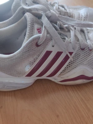 Vita Adidas sneakers med vinröda detaljer - Snygga vita sneakers från Adidas med klassiska tre vinröda ränder på sidan och matchande vinröda detaljer på sulan. Ovandelen är i mesh för bra ventilation och skon har en sportig, låg profil. Perfekta för dig som gillar en clean och sportig look.