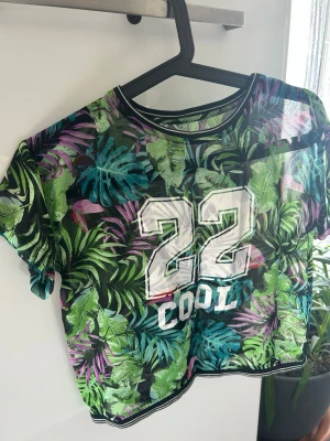 Grön croppad t-shirt med tryck - Säljer en croppad t-shirt med färgglatt blad-mönster i grönt, blått och lila. Framtill står det '22 COOL' i vitt tryck. T-shirten har rund hals och korta ärmar, perfekt för dig som gillar streetstyle och vill sticka ut man kan hälst ha något under eftersom att den är genomskinlig.