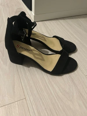 Svarta klackpumps från Seastar - Stilrena svarta pumps från Seastar med öppen tå, bred rem över foten och justerbart spänne vid ankeln. Skorna har en bekväm blockklack och ovandelen är i mockaimitation. Perfekta för dig som vill ha en klassisk och enkel look.