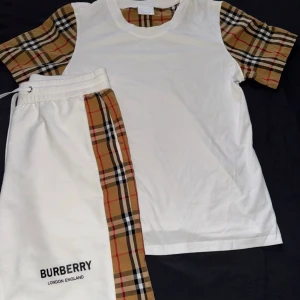 Vita shorts med rutiga detaljer med en vit tröja - Säljer ett par vita shorts och ett par vit T-shirt från med klassiska beige-rutiga paneler längs sidorna och svart logga framtill. Storlek L på shortsen men passar M med, och storlek S på tröjan. 