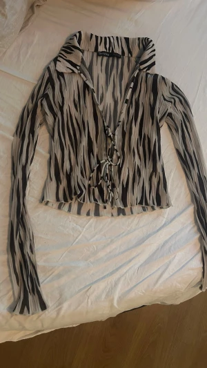 Zebramönstrad blus med knyt framtill - Säljer en zebramönstrad blus från Bershka med långa ärmar och trendiga knyt framtill. Blusen har en krage och är gjord i ett lätt, plisserat material i svart, vitt och beige. Perfekt för dig som gillar statement-plagg och vill sticka ut.