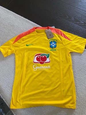 Brasilien fotbollströja och shorts Nike - Säljer ett Brasilien fotbollskit från Nike med gul tröja och svarta shorts. Tröjan har grönt Nike-märke, CBF-logga och Guaraná Antarctica-tryck samt röda detaljer på axlarna. Shortsen är svarta med vit Nike-logga, CBF-märke och röda detaljer på sidorna. Perfekt för träning eller match!