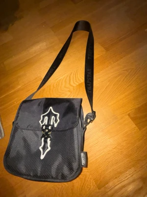 Svart axelväska från Trapstar - Säljer en svart axelväska från Trapstar med justerbar axelrem där loggan är tryckt. Väskan har ett coolt vitt emblem på locket och är gjord i ett slitstarkt mesh-liknande material. Perfekt för att bära det viktigaste och ger en streetwear vibe.