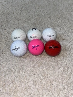 6 golfbollar i olika färger och märken - Säljer ett set med 6 golfbollar från olika märken: Titleist, Top-Flite, Bridgestone, Srixon och Spalding. Bollarna är vita, rosa och röda, alla med klassisk rund form och dimple-mönster. Perfekt för dig som vill ha variation på golfbanan.