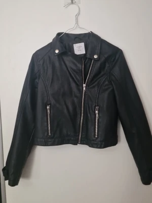 Svart skinnjacka från Lindex Kids 152 - Snygg svart skinnjacka från Lindex Kids med silvriga dragkedjor och tryckknappar. Klassisk bikerstil med bred krage och detaljerade sömmar. Perfekt för en cool och trendig look.