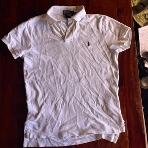 Vit pikétröja från Polo Ralph Lauren - Klassisk vit pikétröja från Polo Ralph Lauren med korta ärmar och blå broderad logga på bröstet. Tröjan har krage och knappar vid halsen. Tillverkad i mjukt bomullsmaterial och har en tidlös, avslappnad passform.
