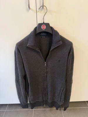 Mörkgrå ziptröja från Polo Ralph Lauren, stl S - Mörkgrå ziptröja från Polo Ralph Lauren med hög krage och diskret logga på bröstet. Tröjan har ribbade muddar vid ärmslut och nederkant samt två fickor framtill. Perfekt för lager på lager och enkel att matcha.