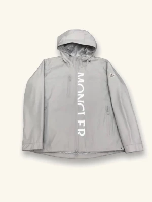 Grå regnjacka från Moncler - Snygg grå regnjacka från Moncler med huva och dragkedja framtill. Stor vit Moncler-logga längs dragkedjan och en diskret logotyp på ärmen. Jackan har fickor med dragkedja och justerbar huva. Perfekt för regniga dagar och streetwear-stil.