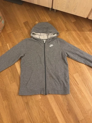 Grå zip-hoodie från Nike  - Grå hoodie från Nike  med dragkedja framtill och två fickor. Klassisk Nike-logga broderad på bröstet. Mjuk insida och huva med ljusgrått foder. Perfekt för en avslappnad och sportig stil.
