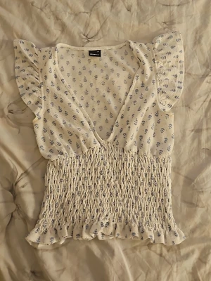 Vit blommig blus med volangärm - Söt vit blus med små blå blommor, v-ringning och volangärmar. Toppen har smockad midja och volangkant nedtill för en feminin look. Perfekt till jeans eller kjol för en fräsch sommarstil.