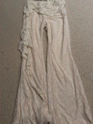 Krämvita spetsleggings med volang - Unika krämvita leggings i spets med utsvängda ben och dekorativ volang längs ena sidan. Materialet är genombrutet med blommönster och ger en romantisk vibe. Perfekta för dig som vill sticka ut med en bohemisk och trendig look.