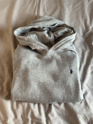 grå ralph lauren hoodie med mörk blått märke - Snygg grå hoodie med huva och känguruficka framtill. Tröjan har en liten broderad logga på bröstet i mörkblått. Tillverkad i mjukt sweatshirtmaterial, perfekt för chill dagar eller när du vill ha en clean och enkel stil.