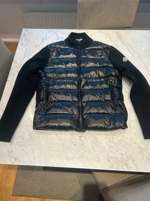 Moncler jacka - Snygg svart dunväst med blanka paneler och stickade svarta ärmar. Västen har dragkedja framtill, ståkrage och två fickor med dragkedja. Moncler-logga på vänster ärm. Perfekt för lager-på-lager och streetstyle.