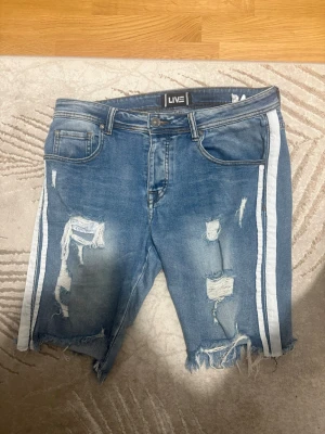 Blå jeansshorts med vita ränder - Säljer ett par blå jeansshorts med slitna detaljer och råa kanter. Shortsen har vita ränder längs sidorna och klassisk femficksdesign. Perfekta för dig som gillar en avslappnad och trendig stil.