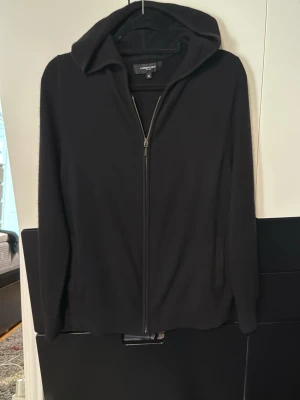 Kashmir hoodie med dragkedja - En stilren svart kashmir hoodie med dragkedja framtill och huva. Hoodien har långa ärmar och två sidofickor. Tillverkad i ett mjukt material som känns skönt mot huden. Perfekt för dig som gillar en clean och enkel look. 100% kashmir