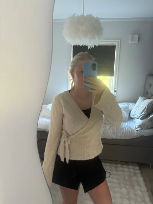 Beige omlottstickad tröja - Säljer en beige stickad omlott-tröja med knytband i midjan. Tröjan har långa ärmar, ribbade muddar och en mjuk, mysig känsla. Perfekt för lager på lager och enkel att styla till olika outfits.