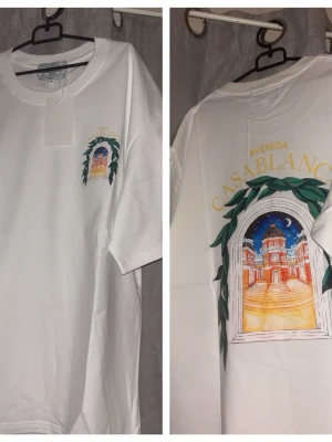 Vit t-shirt med Casablanca-tryck - Vit t-shirt med färgglatt tryck på bröstet och ryggen som föreställer en byggnad och texten 'Avenida Casablanca' i gult. T-shirten har rund hals och korta ärmar. Jätte fin när sommaren är under dess höjdpunkt. 