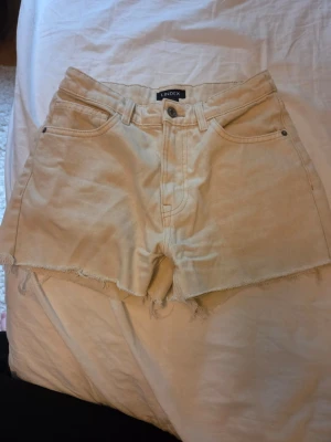 Beige jeansshorts från Lindex - Snygga beige jeansshorts från Lindex med hög midja och råa kanter. Klassisk femficksmodell med knapp och dragkedja framtill. Perfekta för varma dagar och enkel att matcha med olika toppar.