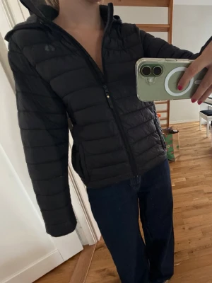 Svart dunjacka med huva - Säljer en svart dunjacka med huva och dragkedja framtill. Jackan har en quiltad design och sitter snyggt figurnära. Perfekt för kyliga dagar och enkel att matcha med olika outfits.
