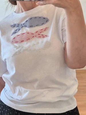 Vit t-shirt med fiskmotiv - Vit t-shirt i bomull med tre fiskar på bröstet i blått, rött och beige rutigt och randigt mönster. Klassisk passform och korta ärmar. Perfekt för dig som gillar lekfulla detaljer och enkel stil.