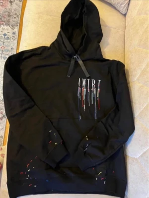 Svart hoodie med färgstänk från Amiri - Snygg svart hoodie från Amiri med huva och dragsko. Framsidan har Amiri-logga med färgstänk i flera färger, och baksidan har en stor logga med samma färgstänk. Perfekt för dig som gillar streetwear och unika detaljer.
