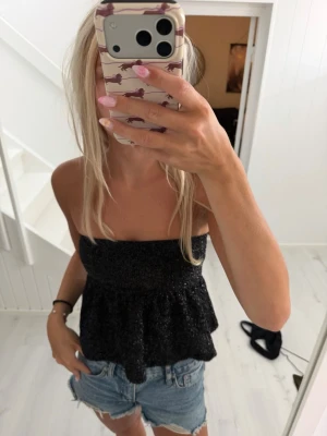 Glittrig svart tubtopp med volang - Säljer en svart glittrig tubtopp med volangkant. Toppen är axelbandslös och har en bandeauformad överdel som sitter tight upptill och faller ut nedtill. Perfekt för dig som vill ha en trendig och snygg topp till sommaren.