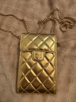 Guldig quiltad handväska med kedja - Supersnygg liten handväska i guld med quiltat mönster och kedjerem. Kedjan kan tas av ifall man vill ha som en clutch bara. Väskan har ett klassiskt lock med tryckknapp och flera fack inuti för kort och småsaker. Perfekt för dig som vill ha en statement-accessoar.