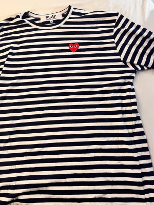 Randig långärmad tröja Cdg PLAY - Säljer en långärmad tröja från PLAY med marinblå och vita ränder samt ett rött hjärta broderat på bröstet. Tröjan har rund hals och är i mjuk bomull, perfekt för en avslappnad och trendig look. Säljer då jag fått den i present och ej passar mig* Bröst: 58 cm * Axel: 53 cm * Ärm: 63 cm * Längd: 77 cm