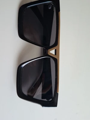 LV Solglasögon  - Snygga solglasögon med bred, svart båge och rektangulära glas. Över bågen löper en guldfärgad metallist med dekorativa detaljer. Glasen är mörka och ger en cool look till alla outfits.