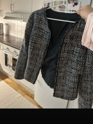 Svartvit tweed cardiganjacka - Stilren svartvit tweed cardiganjacka med rundad halsringning och öppen front. Jackan har ett klassiskt rutmönster och är helfodrad med svart tyg på insidan. Perfekt för att lyfta vilken outfit som helst med en chic touch.