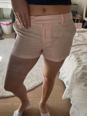 Ljusrosa shorts med bälte - Snygga ljusrosa shorts med midja och bälteshällor. De har en enkel design med fickor på sidorna och en slät baksida. Perfekta för varma dagar och lätta att matcha med olika toppar.