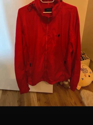 Röd vindjacka från Polo Ralph Lauren - Snygg röd vindjacka från Polo Ralph Lauren med dragkedja framtill, huva och två sidofickor med dragkedja. Jackan har en liten mörk logga på bröstet och justerbar dragsko vid huvan. Perfekt för blåsiga dagar och enkel att matcha.