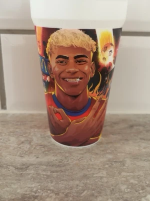 Färgglad plastmugg med illustration - Säljer en färgglad plastmugg med en tecknad illustration av en leende person med blont lockigt hår och färgstark bakgrund. Muggen har motiv i rött, gult, blått och svart och är perfekt för drycker eller som dekoration.