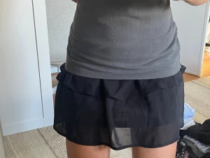 Svart volangkjol i transparent tyg - Säljer en svart kortkjol med volangdetaljer. Kjolen är gjord i ett lätt, transparent tyg som ger en luftig känsla och har en enkel, stilren look. Perfekt för dig som gillar trendiga och feminina plagg.