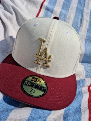 Vit och vinröd LA 59FIFTY keps - Snygg 59FIFTY keps från New Era med LA-logga i guld framtill. Kepsen har vit krona och vinröd skärm, samt broderad MLB-logga bak. Klassisk flat brim och strukturerad passform. Perfekt för dig som vill sticka ut med stilren streetwear.