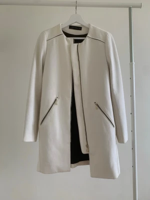 Vit kappa från Zara Woman - Stilren vit kappa från Zara Woman med raka linjer och rund halsringning. Kappan har två snedställda fickor med dragkedjor och stängs med dragkedja framtill. Perfekt för en chic och modern look.