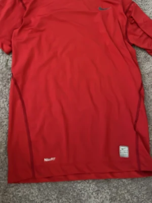 Röd Nike Dri-FIT träningströja - Säljer en röd Nike Dri-FIT träningströja med korta ärmar. Tröjan har en klassisk passform och är tillverkad i ett lätt och snabbtorkande material som passar perfekt för träning. Nike-logga på bröstet och Dri-FIT-märke nedtill.