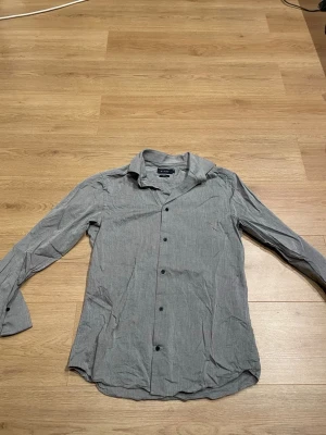 Grå skjorta från Riley slim fit - Snygg grå skjorta från Riley i slim fit-modell. Skjortan har diskret mönster, klassisk krage och svarta knappar framtill. Perfekt för dig som gillar en stilren och enkel look.