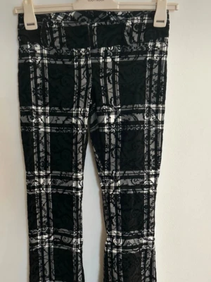 Svarta leggings med rutigt mönster - Snygga svarta flared byxa från BLU FIRENZE med vitt och grått rutigt mönster och dekorativa detaljer. Stretchigt material som sitter tajt och bekvämt. Perfekta för dig som vill ha en unik och trendig look.