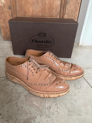 Beige loafers i lack/läder från Church's - Säljer ett par snygga beige lackskor i från Church's i glansigt läder med klassiskt broguemönster och perforerade detaljer. Skorna har en unik ljus sula i kork-look och snörning framtill. Perfekta för dig som vill ha en stilren och tidlös look. Läckra lackskor i beige/brun ton från Church’s. Klassisk modell med riktigt snygg finish. Kanten på sulan har korkdetalj där visst slitage syns, men det påverkar inte funktionen.  Väldigt coola och stilrena skor som lyfter vilken outfit som helst.