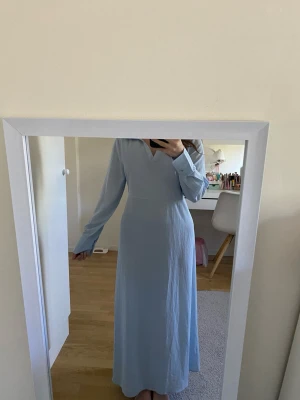 Ljusblå långklänning med knyt i midjan - Snygg ljusblå långklänning med långa ärmar och v-ringad hals. Klänningen har ett elegant knytband i midjan som ger en fin siluett. Perfekt för dig som gillar stilrena och klassiska plagg. Tillverkad i ett mjukt och lätt tyg. Modest. Storleken är S men passar mer Xs