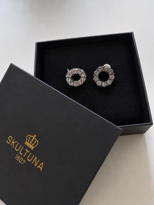 Skultuna runda örhängen med kristaller - Säljer ett par eleganta örhängen från Skultuna i rund form, dekorerade med gnistrande klara kristaller. Örhängena har en silvrig metallbas och kommer i originalask. Perfekta för dig som gillar stilrena och klassiska accessoarer. Lådan medföljer ej men förpackas i smyckesask.