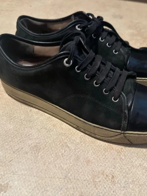 Svarta låga sneakers med gummisula - Säljer ett par svarta sneakers med lågt skaft och klassisk snörning. Ovandelen är i mockaliknande material med tåparti i blankt svart och sula i beige gummi. Perfekta för en clean och stilren look.