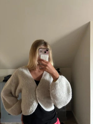 Ljusgrå cropped kofta med ballongärm - Mysig ljusgrå stickad kofta med två knappar framtill och ballongärmar. Koftan är croppad och har en avslappnad passform, perfekt att slänga över en topp. Materialet ser ut att vara mjukt och fluffigt, troligen en ull- eller mohairblandning.