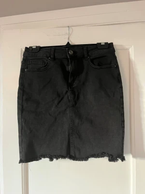 Svart jeanskjol med fransig kant - Säljer en svart jeanskjol med rå, fransig kant nertill och klassiska fem fickor. Kjolen har knapp och dragkedja framtill samt bälteshällor. Perfekt till en avslappnad och trendig look.