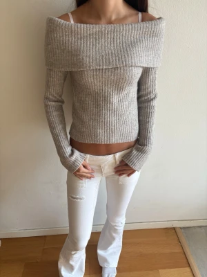 Beige off shoulder stickad tröja - Trendig ribbstickad tröja i beige med off shoulder-skärning och långa ärmar. Tröjan har en bred krage som vilar över axlarna och ger en snygg siluett. Perfekt till jeans eller kjol för en cool look.