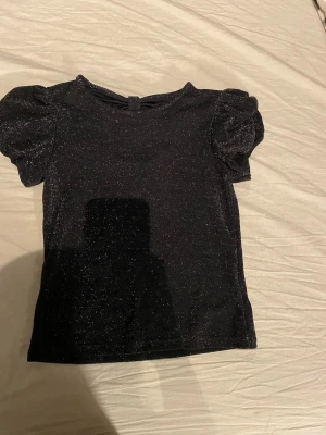 Svart glittrig blus med puffärm - Snygg svart blus med glittereffekt och puffärmar. Blusen har en rund halsringning och en dekorativ knutdetalj med öppning i ryggen. Materialet är stretchigt och skimrande, perfekt för att sticka ut lite extra.