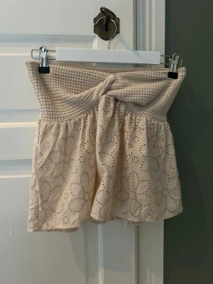 Beige tubtopp med spets och twist - Söt beige tubtopp med twistad detalj framtill och bred smock upptill. Nedre delen är dekorerad med broderad spets i blommönster. Perfekt för sommarens varma dagar. Osäker vart den är ifrån. Har inte kvar lappen med storleken men skulle säga att den är S