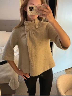 Beige kofta med stora knappar - Snygg beige kofta med trekvartsärmar och hög krage. Koftan har dekorativa stora knappar framtill och ett lätt utställd passform. Perfekt för dig som gillar en stilren och trendig look.